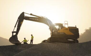 View of two people talking under the boom of a Volvo excavator at dusk with the bucket on the ground.